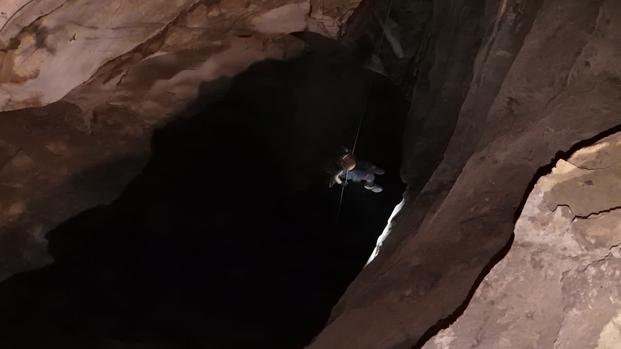 A caver, using Single Rope Technique, descends into a vast, dark opening within Anxiety State Cave, Chiang Mai, Thailand. Their headlamp pierces the enveloping darkness