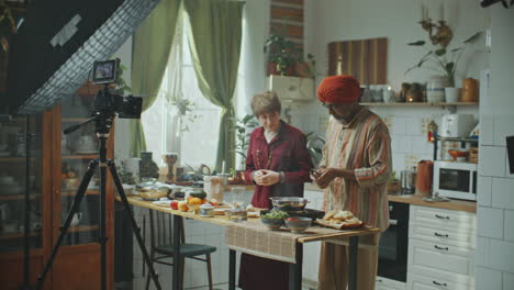 un hombre indio mayor y su co-anfitriona filman un tutorial de cocina en la cocina.