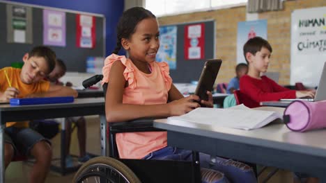 Mixed-race-schoolgirl-in-wheelchair,-sitting-in-classroom-using-tablet,-colleagues-in-background