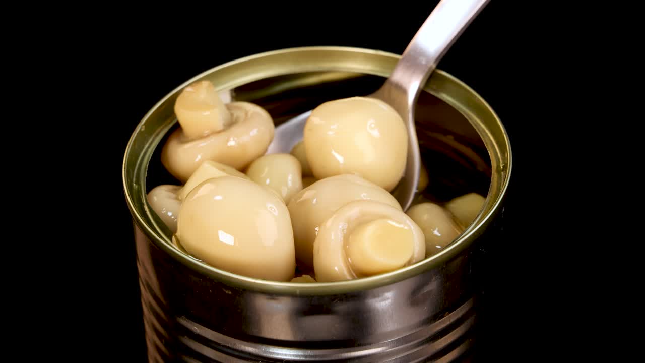 A metal spoon lifts whole champignon mushrooms from an opened can against a black background, with even studio lighting and smooth camera movement