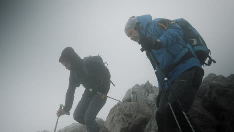 two hikers deciding what direction to take, climbing down from the rocks, cloudy and foggy weather with a strong wind