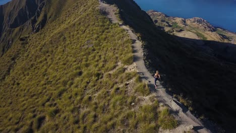 tracking aerial shot of a tall blonde woman running along a trail to the top of a mountain and then stopping and gaining her balance