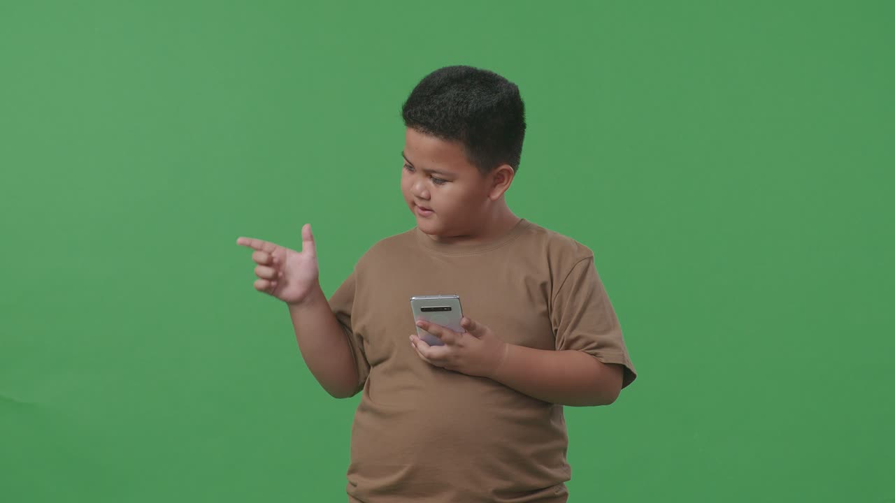 아시아 소년 은 휴대 전화 를 사용 하면서 미소 짓고 스튜디오 의 초록색 화면 배경 에  을 가리키고 있다