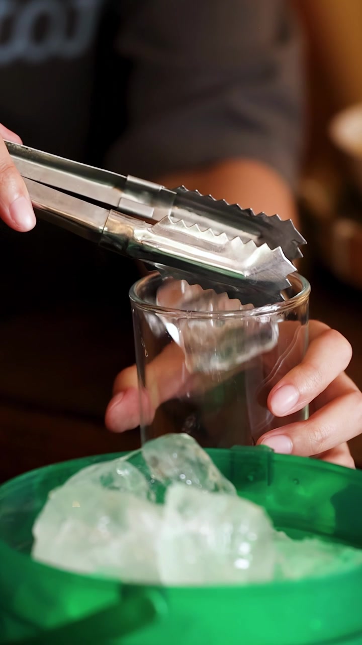 Primo piano di mani che usano pinze di metallo per trasferire il ghiaccio in un bicchiere in Thailandia.