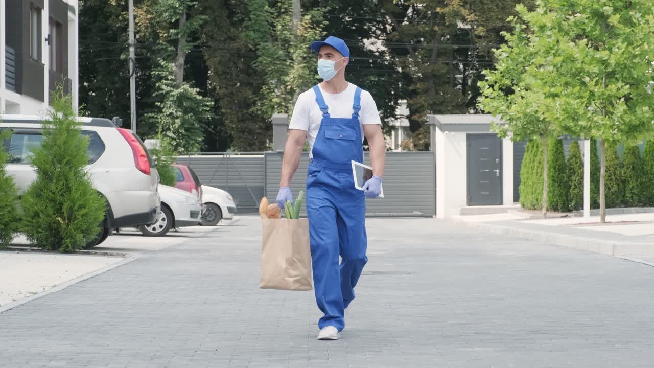 保護マスクと手袋を着用した若い配達員が、食料品の荷物を顧客の自宅へ安全に直接届けます。食品配達、隔離中の宅配。