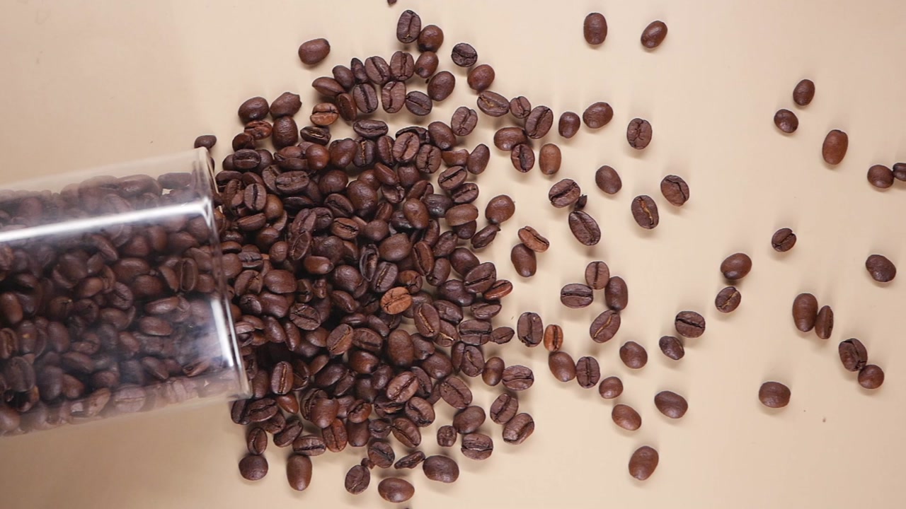 Roasted Coffee Beans Spilling from a Glass Jar on Beige Background