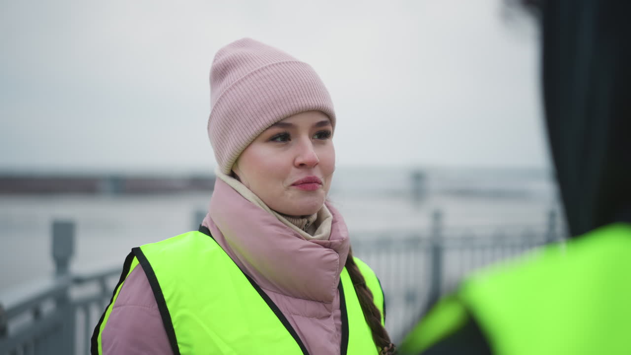 Smiling young woman in pink winter jacket and beanie wearing bright reflective safety vest outdoors during cold overcast day, standing near coworker at industrial site