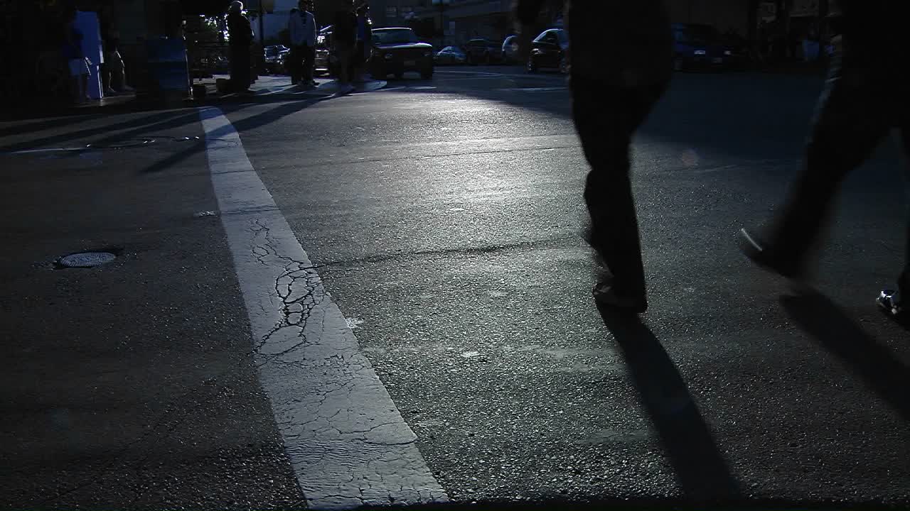 포장 도로를 따라 긴 그림자가 늘어서 보행자가 바쁜 도시 거리의 횡단보도를 통과합니다.