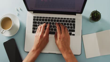 Hands-using-laptop-with-smartphone,-notepad,-plant-and-coffee-on-blue-desk