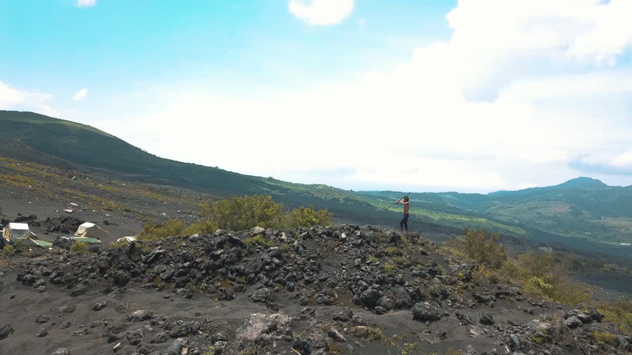 과테말라 파카야의 화산 풍경, 캠핑 장소 근처의 바위에 서서 지팡이를 돌리는 남자의 드론 공중 회전 샷