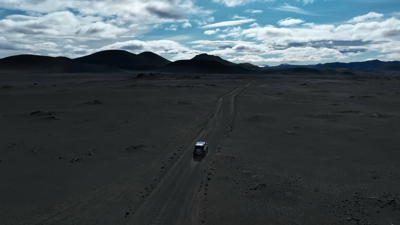 A lone 4x4 drives across Iceland’s black volcanic desert under bright clouds, crossing the remote Fjallabak wilderness with mountains on the horizon