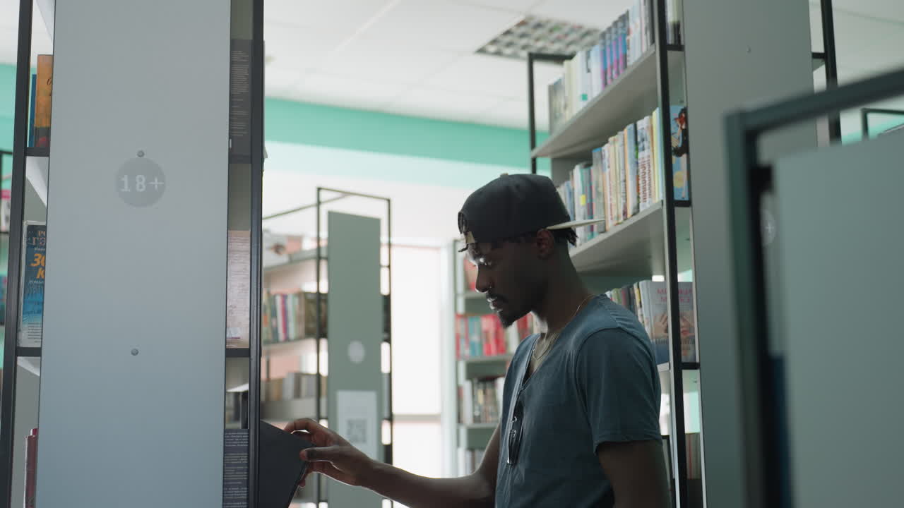 High school student in casual cap and t shirt standing in library aisle, reaching for book on shelf, surrounded by rows of colorful books under bright daylight