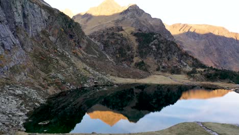 Lac-d'Espingo-lake-reflecting-nearby-peaks-with-calm-water-in-Haute-Garonne,-Pyrénées-mountains,-France,-Aerial-low-flyover-shot