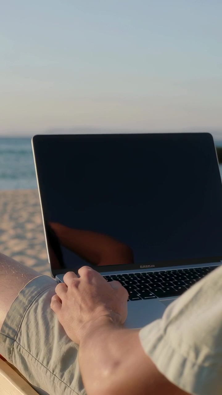 Video verticale: Spostamento della telecamera, uomo che digita al portatile in spiaggia, riflesso dello schermo, lavoro da remoto