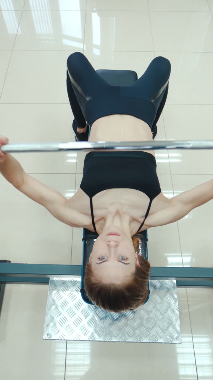 Athletic woman is lying and doing the bench press using the barbell on a white background in the gym. View from above. Endurance exercise Vertical video