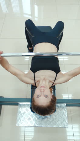athletic woman is lying and doing the bench press using the barbell on a white background in the gym. view from above. endurance exercise vertical video