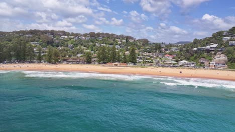 Luftaufnahme-In-Richtung-Surfer-Und-Den-Strand-Gebäude-Und-Häuser-Auf-Den-Klippen-Dahinter