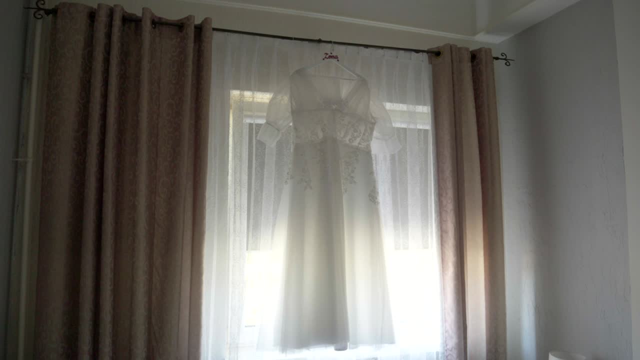 White Wedding Dress Hanging on Curtain Rod by Bright Window