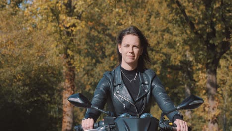 vista conmovedora de una joven europea bastante sonriente conduciendo una moto con chaqueta de cuero en el bosque con hojas de otoño doradas vibrantes y coloridas en un día soleado