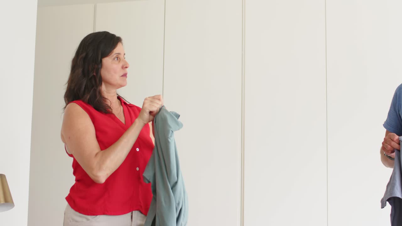 Coppia che sistema vestiti da donare sul letto, la donna piega una maglietta verde in una scatola, con spazio per il testo.