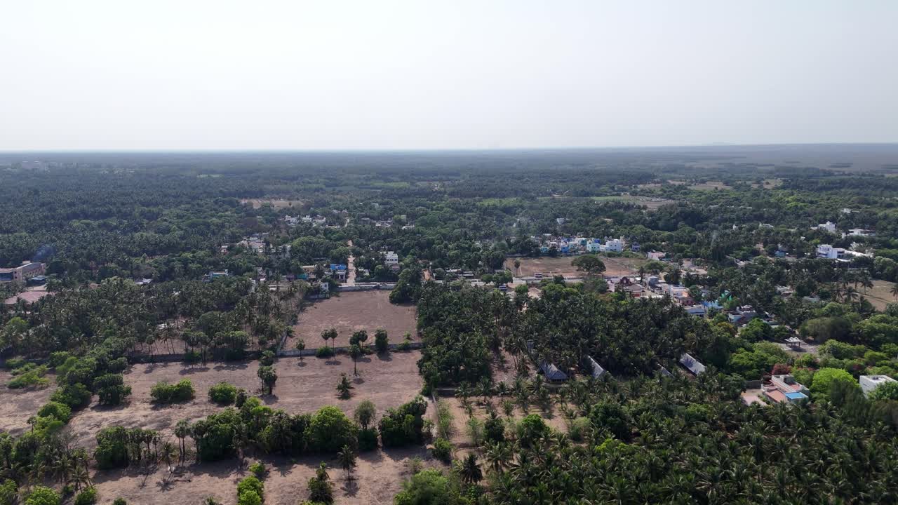 Aerial drone shot of a tranquil rural coastline in South India. A sandy beach meets a dense forest of palm trees and scattered houses, with a cleared patch of land