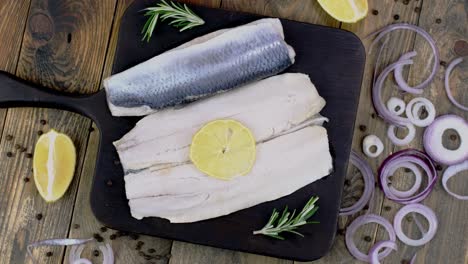 fillet of salted herring with onions, lemon and rosemary on dark cutting board rotating.