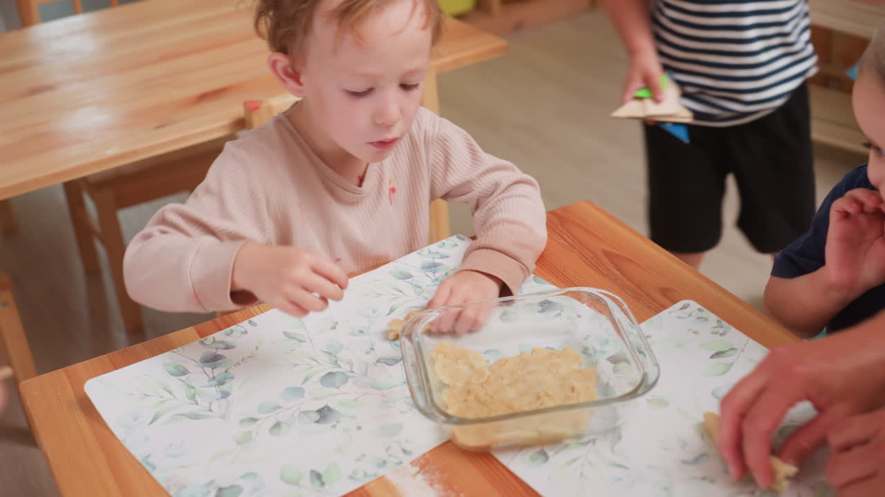 Bambino che impara a modellare l'impasto all'interno di una teglia di vetro mentre un altro bambino si avvicina con in mano una forma di pasta ritagliata, dimostrando concentrazione, capacità motorie, lavoro di team e attività interattiva di cucina