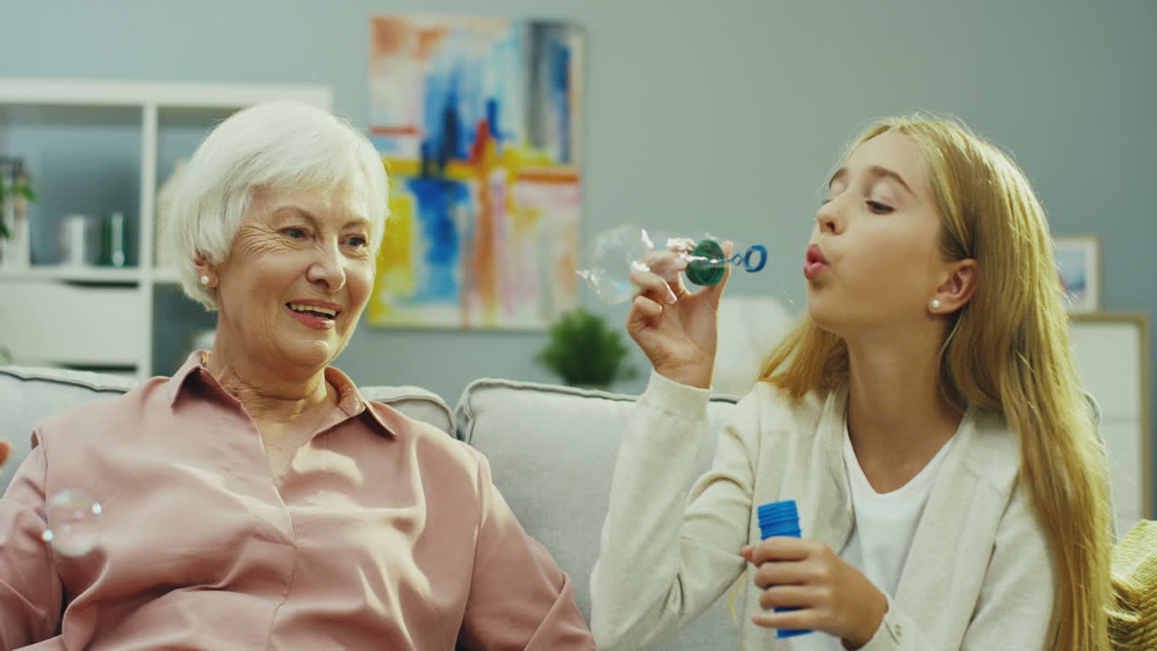 길고 밝은 머리카락을 가진 귀여운 소녀가 집에서 소파에 행복한 할머니와 함께 앉아서 비누 거품을 불어넣고 있습니다. 실내에서. 초상화 촬영.