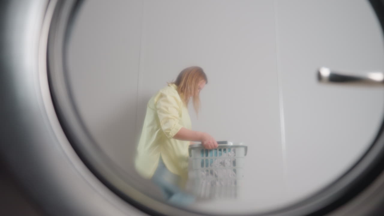 Washer door reflection shows homemaker with laundry basket dancing backward with smile, playful pass across frame, round window rim and handle visible, bright interior, joyful chore moment