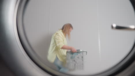 washer door reflection shows homemaker with laundry basket dancing backward with smile, playful pass across frame, round window rim and handle visible, bright interior, joyful chore moment