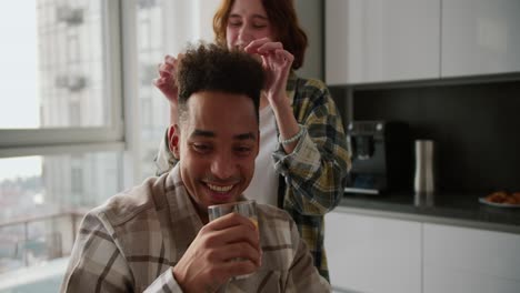 Portrait-of-a-cheerful-and-happy-young-man-with-Black-brunette-skin-color-in-a-checkered-beige-shirt-who-sits-and-drinks-juice-while-his-girlfriend-touches-his-hairstyle-in-a-modern-apartment