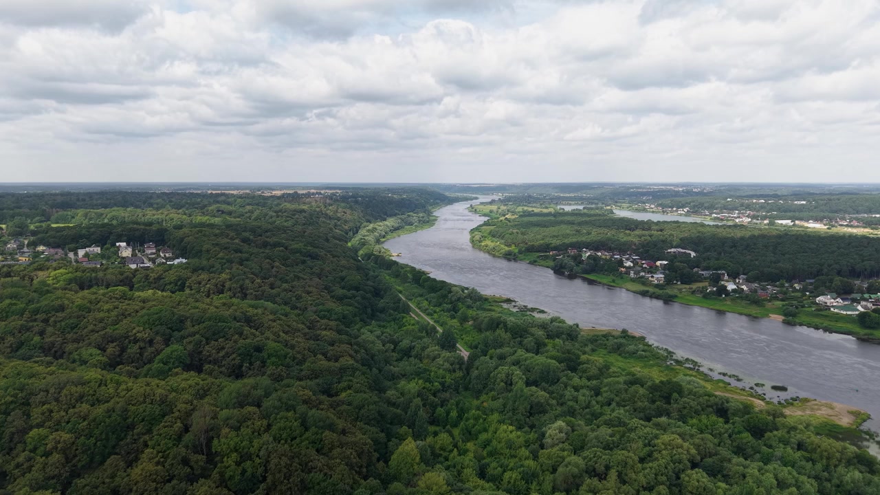 Video drone che sorvola un fiume ampio e sinuoso delimitato da una fitta foresta verde e villaggi lontani, rivelando un vibrante fogliame estivo e un paesaggio naturale pacifico in una luce diurna soffusa.