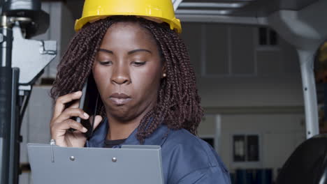 close up of an african american female worker talking on phone, standing in storehouse and holding a clipboard