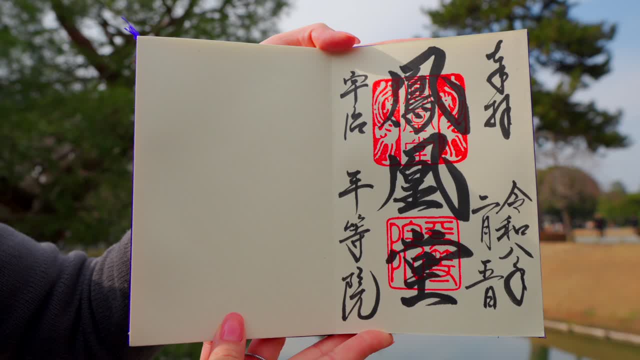Una donna bianca tiene un Goshuincho, un album speciale per timbri con un timbro Goshuin fatto a mano, presso lo storico Tempio Byodo-in a Uji, Giappone. Inquadratura statica.