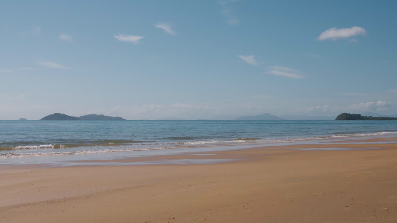 僻静的海滩，轻柔的海浪拍打着岸边，背景是岛屿，蓝天中飘浮着洁白的朵朵浮云。