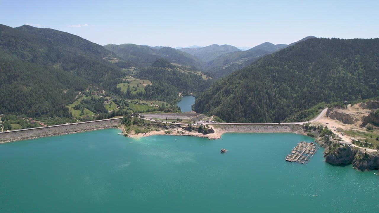 An aerial drone reveals Zaovine Lake in Serbia, its long dam and a floating fish farm below a broad mountain valley, with forested slopes, rural roads and rocky shore in daylight.