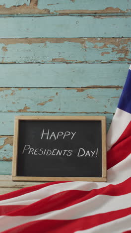 Vertical video of happy presidents day text on chalkboard with american flag against painted boards