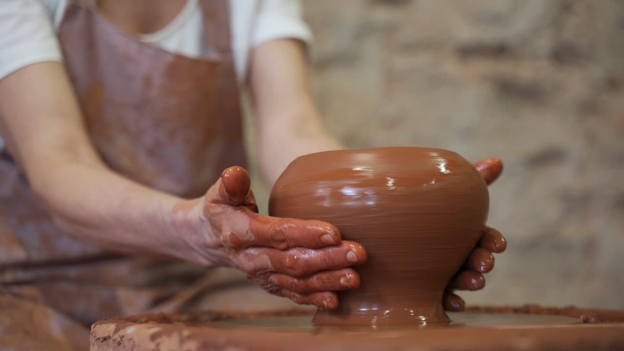 el escultor en el estudio hace un primer plano de una olla de arcilla. una mujer mayor está esculpiendo un cuenco detrás de una rueda de alfarero giratoria