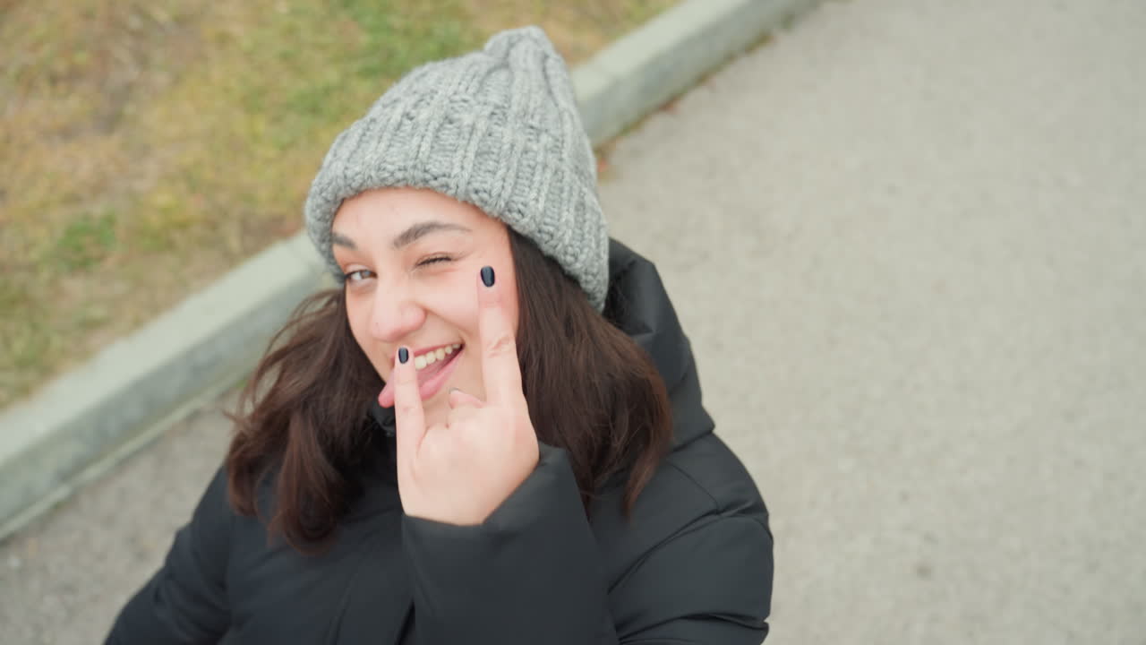 Overhead woman walking pavement playful dance moment, street portrait of woman in grey knit beanie and black puffer coat walking along curb, expressive smile and spontaneous finger touch to cheek