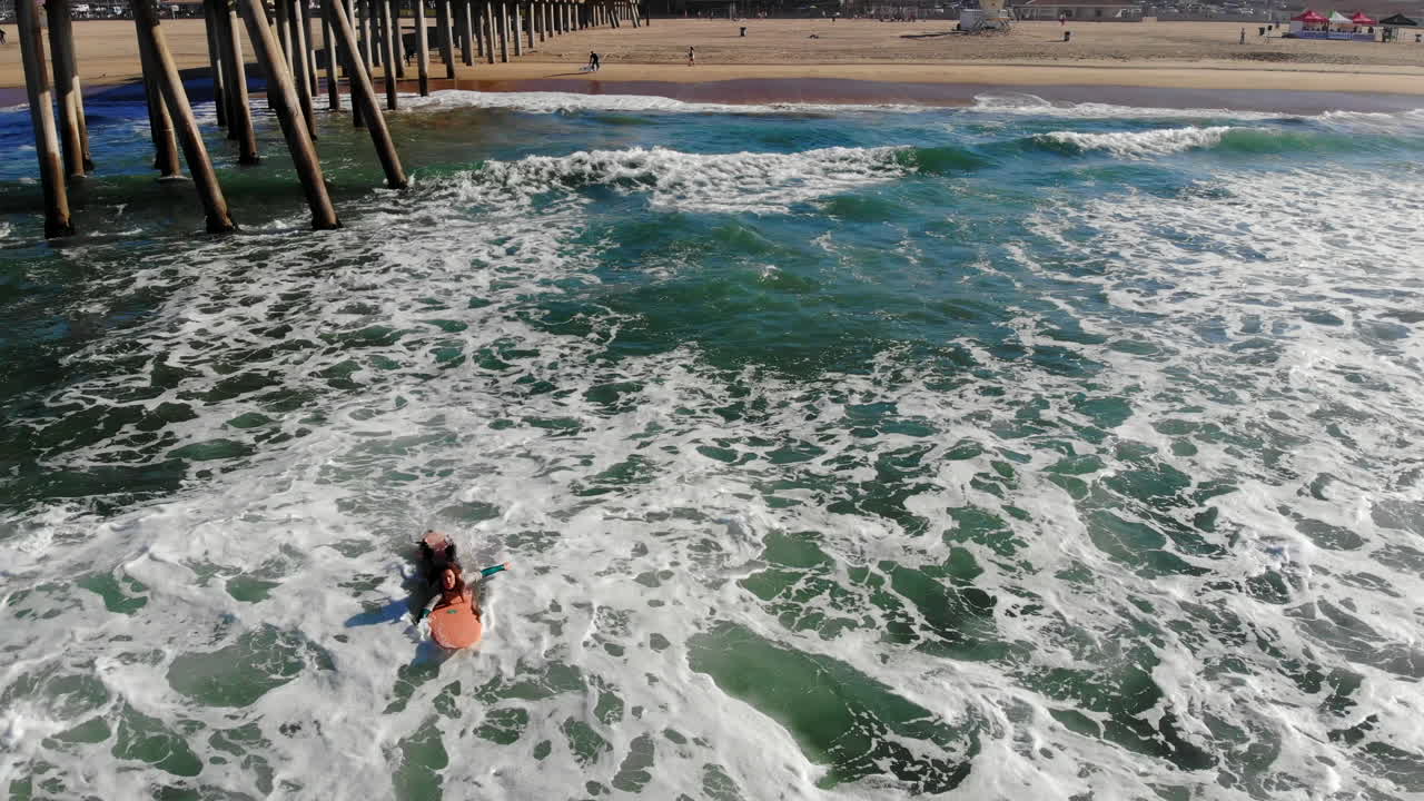 una surfista rema hacia las olas en el muelle de la playa de huntington en una competencia de surf con un dron aéreo de 4k volando hacia atrás sobre las olas del océano pacífico a primera hora de la mañana en el sur de california