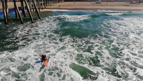 Female-surfer-paddles-out-into-waves-at-Huntington-Beach-Pier-in-surfing-competition-with-4k-aerial-drone-flying-backwards-over-Pacific-Ocean-waves-in-early-morning-in-Southern-California