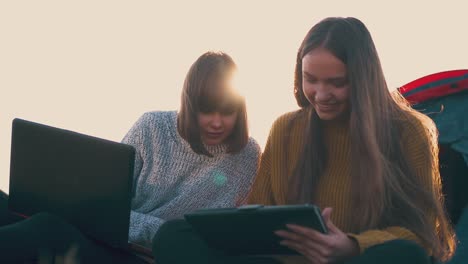 girl-shows-tablet-info-to-friend-with-laptop-by-tent-in-camp