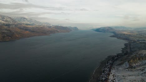 Eine-Stimmungsvolle-Weitwinkelaufnahme-Des-Kamloops-sees-Im-Winter,-Umgeben-Von-Einer-Hügeligen-Wüstenlandschaft,-Die-Teilweise-Mit-Schnee-Bedeckt-Ist,-Und-Einem-Zug,-Der-An-Seinen-Stränden-Entlang-Fährt