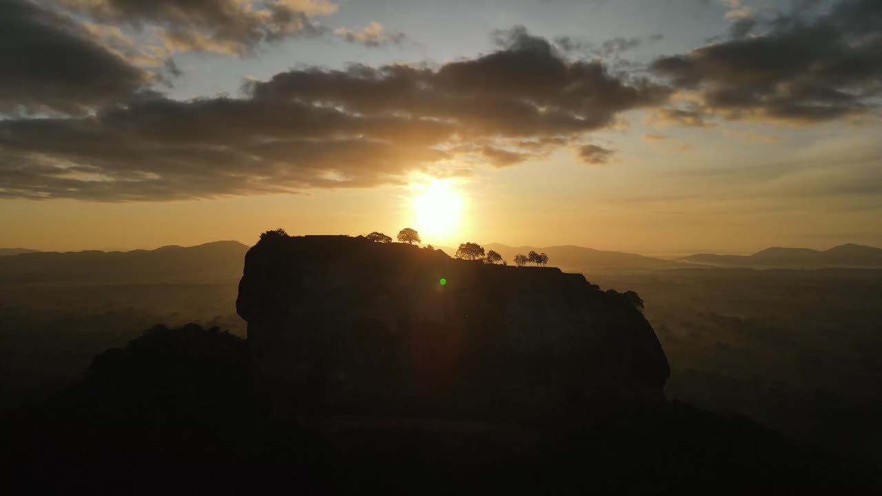 Beautiful yellow sun setting behind the silhouettes of the trees of the citadel called Sigiriya in Sri Lanka with in de background the big mountains on a cloudy day. Wide drone lowering dolley shot