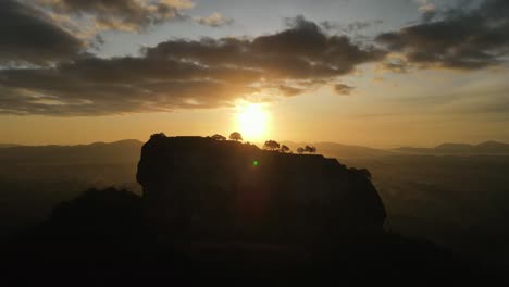 beautiful yellow sun setting behind the silhouettes of the trees of the citadel called sigiriya in sri lanka with in de background the big mountains on a cloudy day. wide drone lowering dolley shot