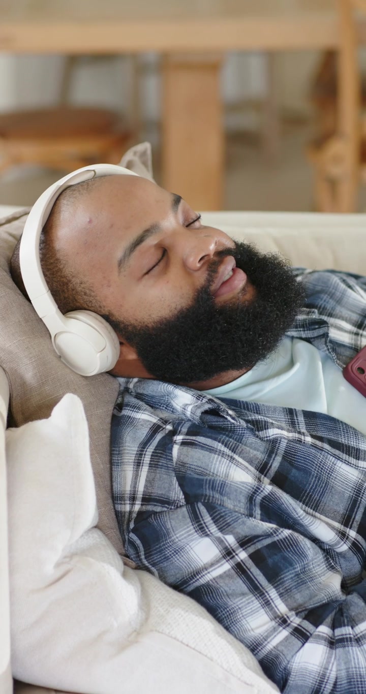 Afroamerikansk mand læner sig tilbage på sofaen, lytter med hovedtelefoner, telefonen afspiller musik for at slappe af