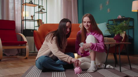 girls friends siblings sitting on floor and take turns dropping dollar banknote into piggy bank