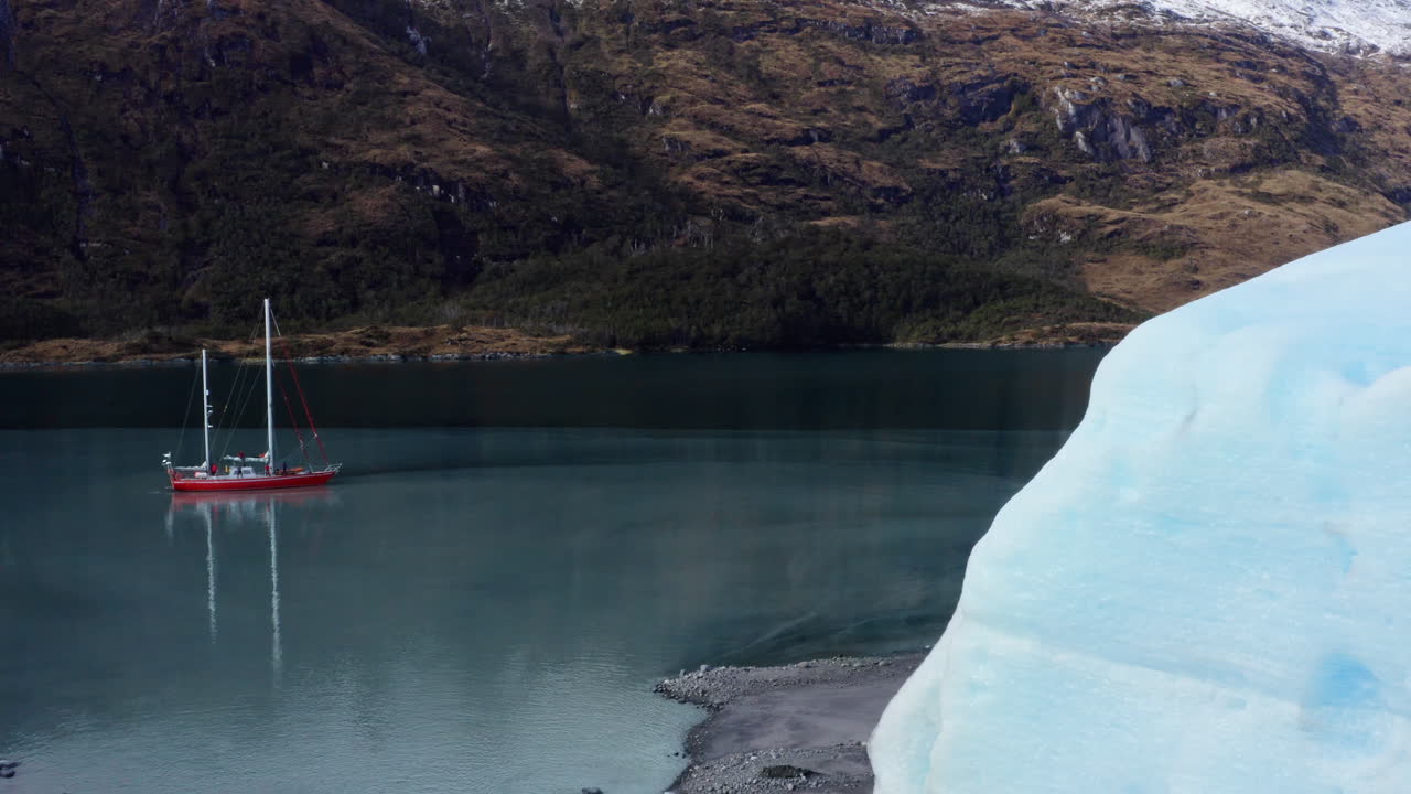 Iceberg floats in cold Patagonian waters with visible edge detail and reflection in sea, aerial reveals sailboat in turquoise green water