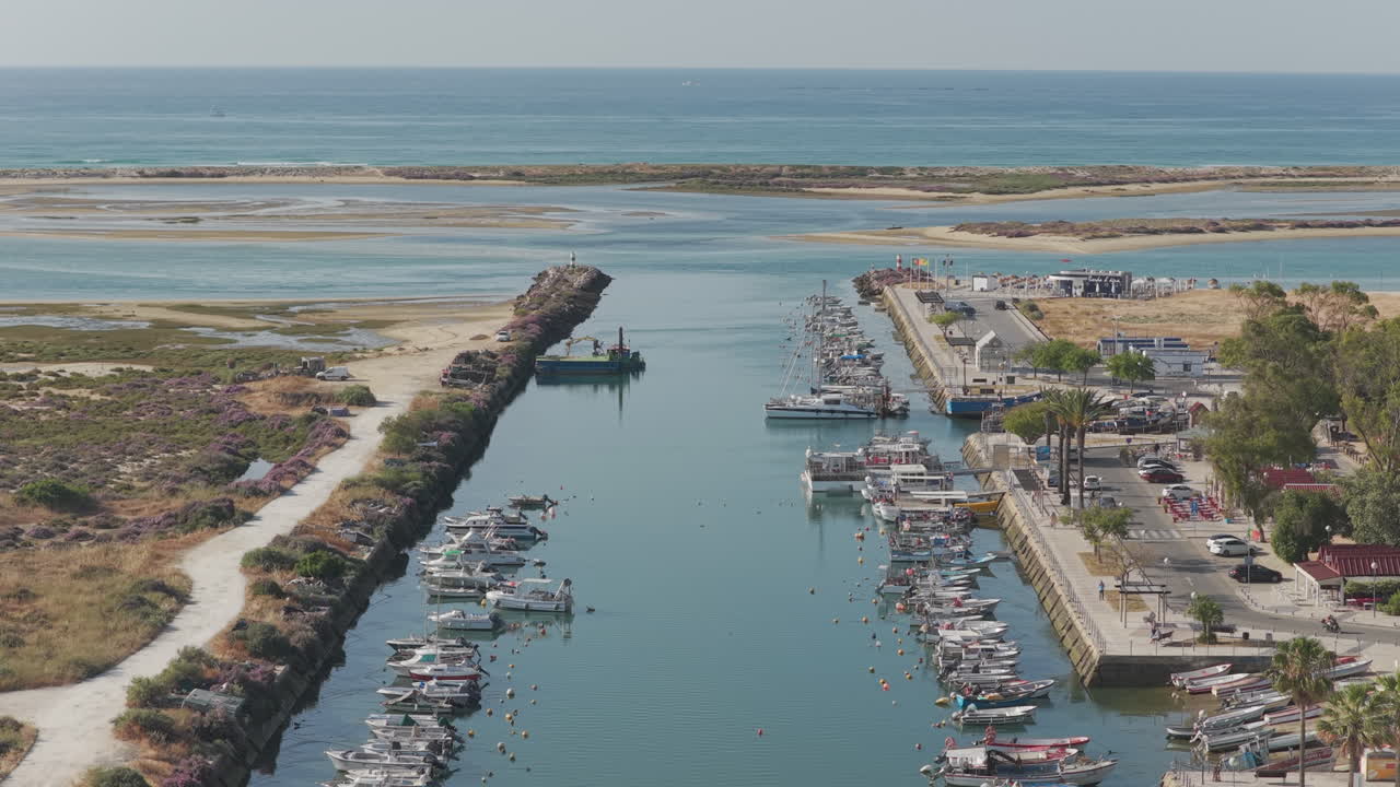 Drone flying forward above Fuseta harbour channel, showing boats moored on both sides, sandy dunes on the left and port buildings on the right, with the ocean visible in the distance