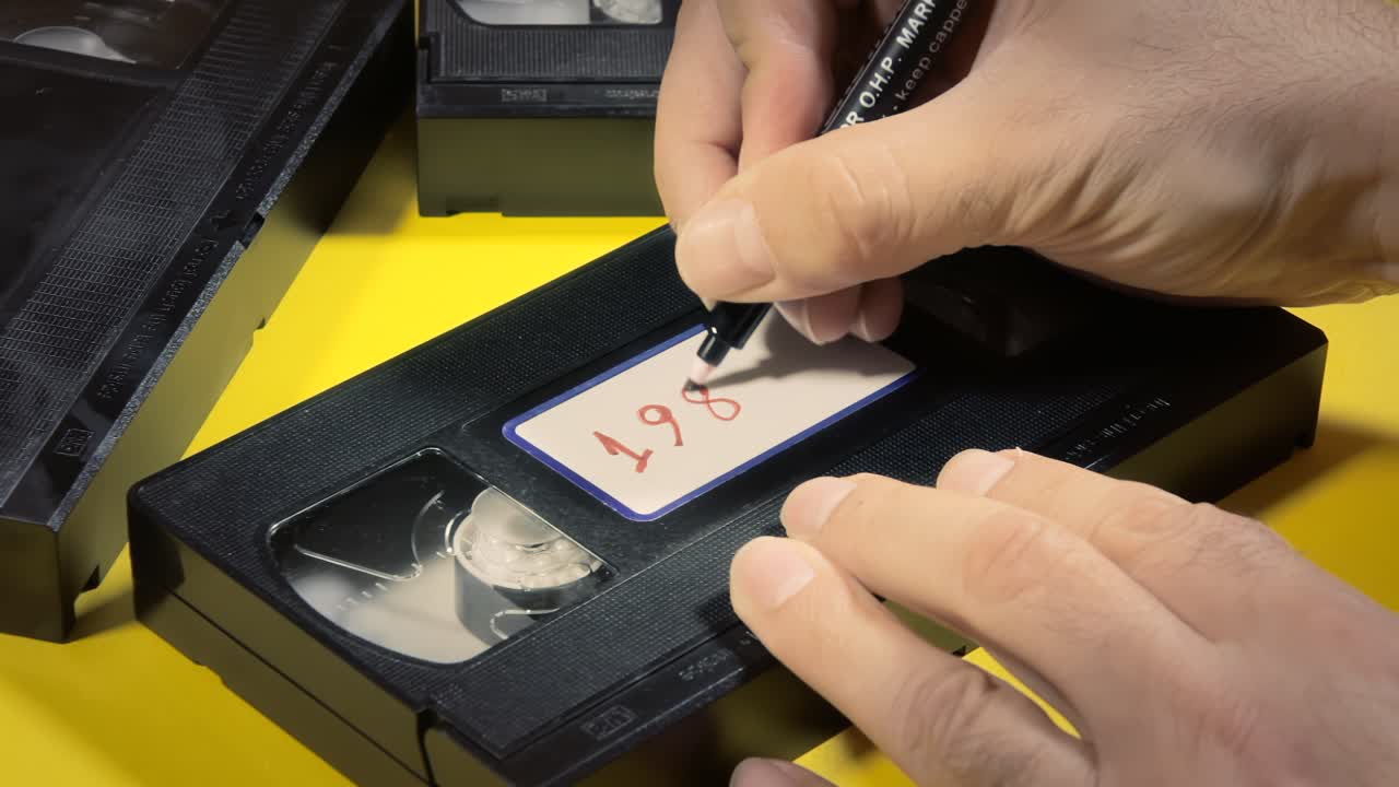 Writing the text 1980's on the white label of an old VHS videotape , with a marker
